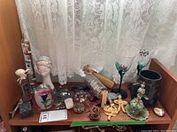 Wide view of shelf with assorted decorative items including glass bottle with flower stopper, Galileo thermometer, ceramic bust, crosses, glass paperweight, ship in bottle, metal coasters, and other small items.