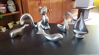 Group photo of all five Hoselton brushed aluminum figurines showing polar bear, fox, whale, swan, and sailboat on a table with a mid-century modern wooden cabinet in the background.