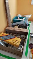 Photo showing assorted metal baking molds, tart pans, icing tools, knives and peelers in boxes and containers.