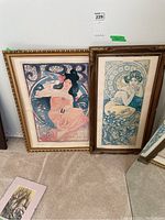 Pair of framed Alphonse Mucha Art Deco era prints leaning on carpeted floor showing full view of both prints and gold-tone frames