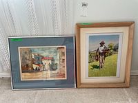 Two framed original paintings side by side against a wall, one with blue mat and glass, one in wood frame.