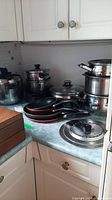 Seven assorted pots, four fry pans stacked, one roasting pan, steamer pot, and steam top, all placed on counter.