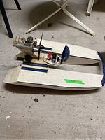 White and blue remote controlled catamaran boat on floor with exposed 0.5 engine and propeller, showing top and side views.