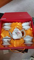 Full view of boxed tea set with one teapot in center and four cups with saucers arranged in original box lined with yellow satin.