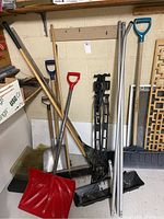 Full view of all tools leaned against a wall including brooms, shovels, rake and snow grabber