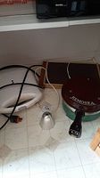 Photo showing the Remoska electric pot with maroon lid and handle alongside the warming tray and additional small metal accessory with power cord visible.