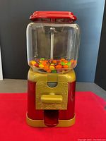 Front view of the candy vending machine filled with Skittles for display. Shows the red metal base, gold coin mechanism, clear glass container with carousel inside, and red lid on top.