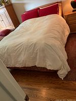 White down duvet shown laid out on a queen size bed in a bedroom setting, visible red pillows and nightstand lamp.