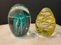 Two colorful glass art paper weights showing jellyfish and yellow abstract designs, placed side by side on white cloth background