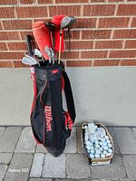 Wilson golf carry bag filled with golf clubs, beside box of used golf balls on pavement with brick wall background