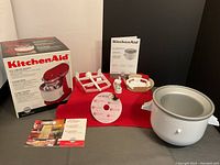 Full lot overview showing original KitchenAid box, ice cream maker components arranged on grey surface with red cloth underneath, and user manuals.