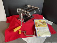 Side view of the Marcato manual pasta maker with detachable crank handle and clamp, placed on red and grey cloths.