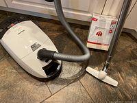 White Miele canister vacuum with gray hose and hardwood floor attachment beside sealed filter box on tile floor
