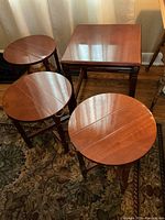 Four wooden nesting and folding side tables shown together on a patterned rug in front of a curtain.