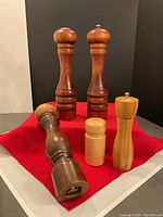 Front view of all five items arranged on red cloth: two medium brown pepper mills with silver knobs, tall dark brown curved pepper mill, light wood salt shaker, light wood pepper grinder.