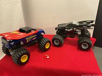 Photo showing both Superman (blue, yellow wheels) and Batman (black, gray wheels) monster trucks, plus the small red Marvel pin in front on red cloth