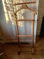 Full front view of the vintage wooden valet showing its hanger, trouser bar, and wheels at the base.