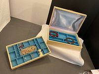 Full view of open vintage cream-colored jewellery box with blue felt lined removable tray with separated compartments containing jewellery. Box interior lid is satin. Contains bracelets, earrings, pendants, and necklace. Beige case exterior shows age wear.