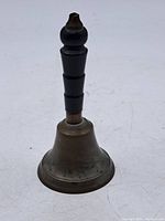 Metal antique school bell with dark wooden turned handle standing upright, showing the exterior with signs of wear and tarnish.
