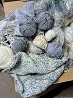 Box of assorted yarn balls and skeins in light blues, greys and white tones, showing mixed textures and knit sample.