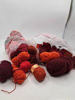 Overall view of multiple skeins and balls of yarn in reds and oranges spilling out of plastic bags on white background. Various shades and thicknesses visible.