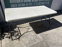 White folding plastic table set up outdoors on pavement, showing full table surface and metal legs.