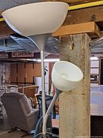 Floor lamp showing two light fixtures: a large upward-facing bowl lamp and a smaller adjustable lamp, set against a wood post in a basement or workshop setting.