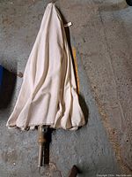 Beige fabric patio umbrella folded with attached brown metal pole laying on concrete floor.