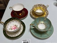 Four different fine china tea cup and saucer sets grouped together to show variety, colors, and gold decorative patterns.