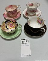 Overview photo of all four china tea cups and saucers, showing varied floral patterns and colors including pink, black, white, and yellow with gold accents.