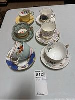 Overall view of seven fine china tea cups and saucers arranged on table showing different patterns and colors, highlighting mixed matching and non-matching pairs.