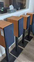 Four wooden veneer speakers with black front grills on black metal stands arranged in a row.