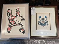 Two framed Indigenous art prints side by side, one in silver frame with red and black abstract figure, the other in brown frame with blue and black frog design.
