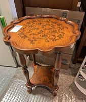 Side angle view of two-tier wooden side table, showing scalloped edges, turned legs, lower shelf with floral inlay, and curved stretcher base.