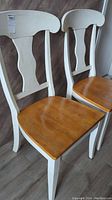 Photo showing a close-up of the two wooden chairs with white backs and natural wood seats, side by side against a wooden wall.
