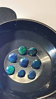 Eight faceted black Ethiopian opal gemstones in a dark dish, showing brilliant rainbow flashes of green and blue.