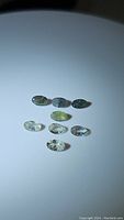 Eight natural alexandrite gemstones placed on white surface, showing difference in color from dark opaque to light off-white stones with facets and inclusions visible.