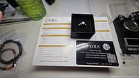 Top down photo showing the ring in its box on a GRA certificate for the moissanite stone and a GRA warranty card.