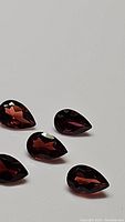 Close-up photo showing five pear cut cherry red garnet gemstones arranged on white background. The gems exhibit bright, deep red color and facet reflections.