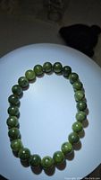 Close-up photo of the bracelet arranged in a circle on a white plate on a dark surface, showing polished round green jade beads evenly strung.