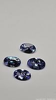 Four oval cut purple-blue tanzanite gemstones with high clarity and luster photographed on a neutral background.