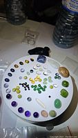 Full view of assorted polished gemstone cabochons on white circular tray showing variety of colors and shapes including deep purple, green, black, and multicolored stones