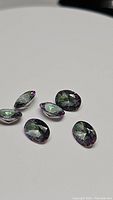 Six oval cut Mystic Topaz gemstones laid on a white surface, showing their purple and multicolor rainbow sparkle.