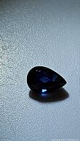 Close-up photo of the natural pear cut blue sapphire gemstone showing its deep blue color and shape against a textured surface.