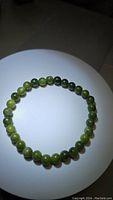 Photo showing circular green jade bead bracelet with varying green shades and reflective polished surface, placed on a white circular platform, well lit to highlight bead color and luster.