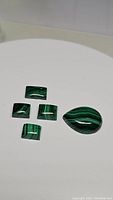 Five polished natural malachite stones arranged on a white background showing rich green banded patterns and various shapes.