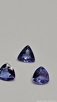 Three natural trillion cut tanzanite gemstones arranged on a gray background showing their purple-blue color and clarity.