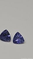 Trillion cut natural Tanzanite pair displayed on a neutral background showing purple blue color and inclusions.