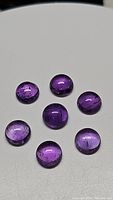 Seven round deep purple amethyst cabochons with smooth dome surfaces showing natural inclusions, arranged on a white background.