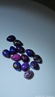 Photo of 12 polished purple copper turquoise gemstones in various shades of purple, blue and copper with natural patterns.